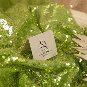 May include: A small, brown card with the text "SECRET SPARK fabrics" and a stylized "SS" logo. The card is lying on a green sequined fabric.