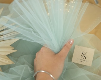 Może przedstawiać: Ręka trzyma jasnozielony tiul z brązową kartką, na której widnieje napis "SECRET SPARK fabrics" czarnym kolorem. Tiul jest złożony w kształt wachlarza.