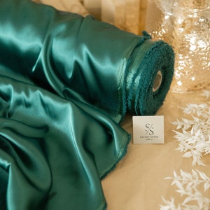 Puede incluir: Un rollo de tela de sat&eacute;n de color verde azulado con un borde con flecos. La tela se muestra sobre una superficie marr&oacute;n con una peque&ntilde;a etiqueta blanca que dice "SECRET SPARK fabrics".