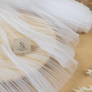 May include: White tulle fabric with a pleated texture. The fabric is folded and arranged on a brown surface. The Secret Spark Fabrics logo is visible on a small brown tag.