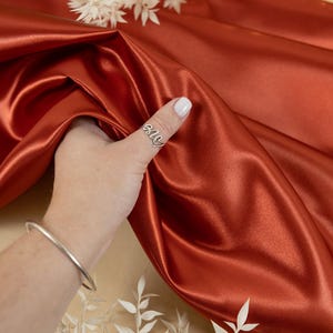 May include: A close-up of a rust-colored satin fabric. The fabric is draped over a hand wearing a silver ring with the word "ally" on it. The hand is also wearing a silver bangle bracelet.