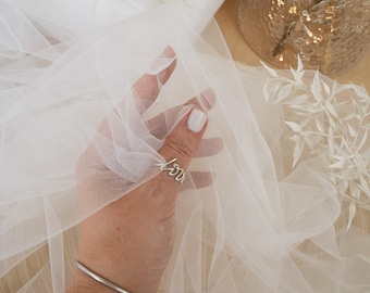 May include: White tulle fabric with a delicate texture. The fabric is draped over a hand wearing a silver ring with the word "love" inscribed on it. The fabric is also draped over a wooden surface.