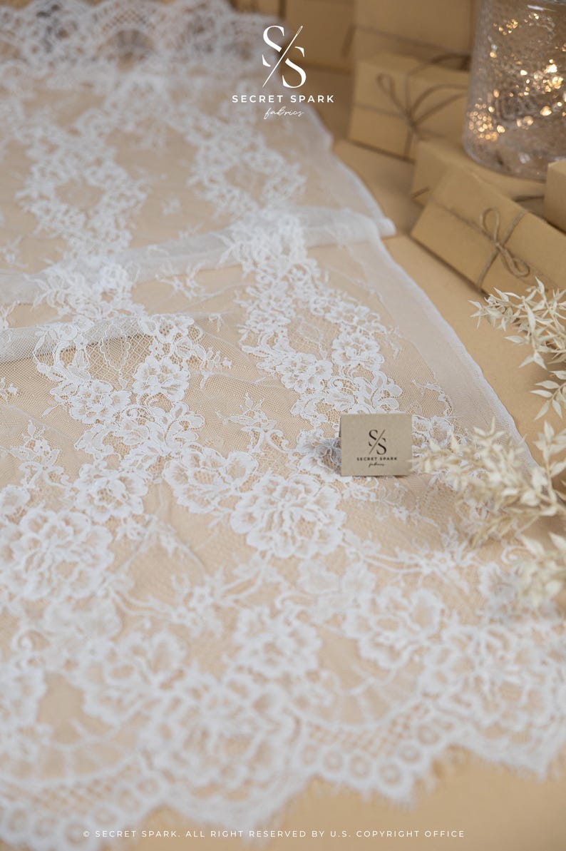 May include: White floral lace fabric with a delicate pattern. The fabric is spread out on a neutral surface, with a small card bearing the "Secret Spark fabrics" logo. The overall aesthetic is soft and elegant.