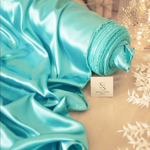 Op de afbeelding: Een rol turquoise satijnen stof met een wit label waarop "SECRET SPARK fabrics" staat.