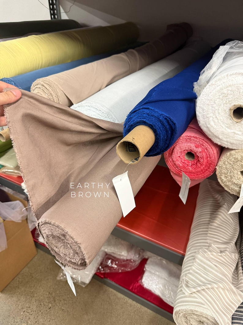 May include: A roll of earthy brown fabric displayed on a shelf alongside various other textile rolls in colors including yellow, blue, white, pink, and striped patterns. The brown fabric roll features a white tag with the text EARTHY BROWN printed on it.