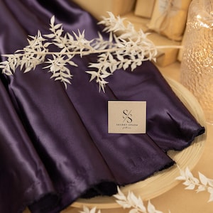 Puede incluir: Primer plano de una tela de satén de color morado oscuro con una etiqueta marrón que dice "SECRET SPARK fabrics".