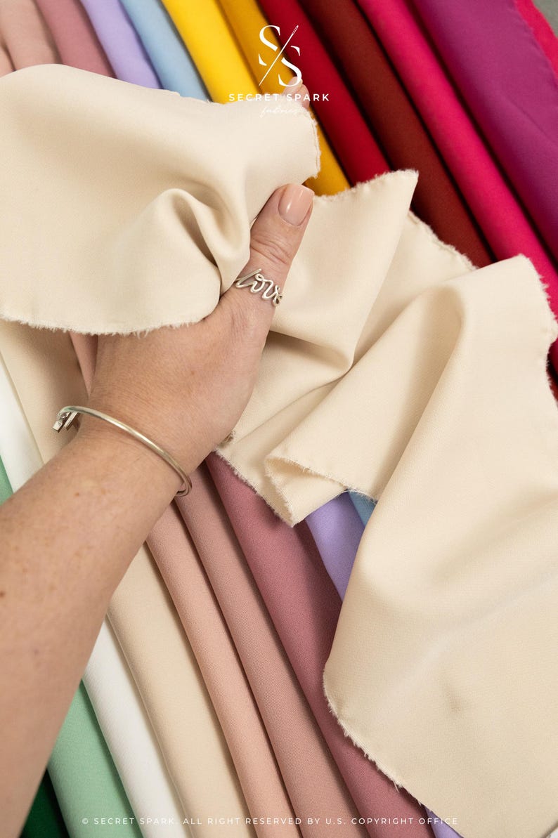 May include: A collection of fabric swatches in various colors, including beige, light pink, lavender, and green. The fabric is smooth and appears to be a lightweight material. The swatches are arranged in a stack, with the beige swatch on top. The text "SECRET SPARK" is visible in the upper right corner of the image.
