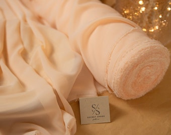 May include: A roll of peach-colored chiffon fabric with a frayed edge. The fabric is rolled up and sitting on a brown surface. A small, brown tag with the text "SECRET SPARK fabrics" is visible in the foreground.