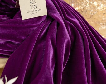 May include: A close-up of a deep purple velvet fabric. The fabric is draped and folded, creating soft folds and wrinkles. The fabric is smooth and luxurious, with a subtle sheen. The fabric is labeled with a small tag that reads "SECRET SPARK fabrics".