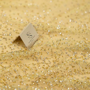 May include: A close-up shot of a golden fabric, densely covered with silver and gold sequins. A small tag with the "SECRET SPARK fabrics" logo is visible. The fabric has a fine, net-like weave, suitable for evening wear or bridal designs.
