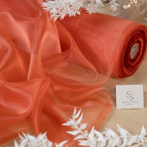 May include: A roll of orange sheer fabric with a subtle shimmer. The fabric is draped over a brown surface with white branches.