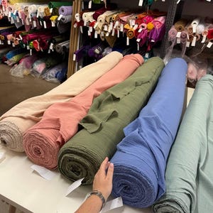 May include: Five large rolls of textured fabric in beige, coral, olive green, blue, and sage green are arranged on a white table. Behind the table, metal shelving units are filled with numerous additional rolls of fabric in various colors.