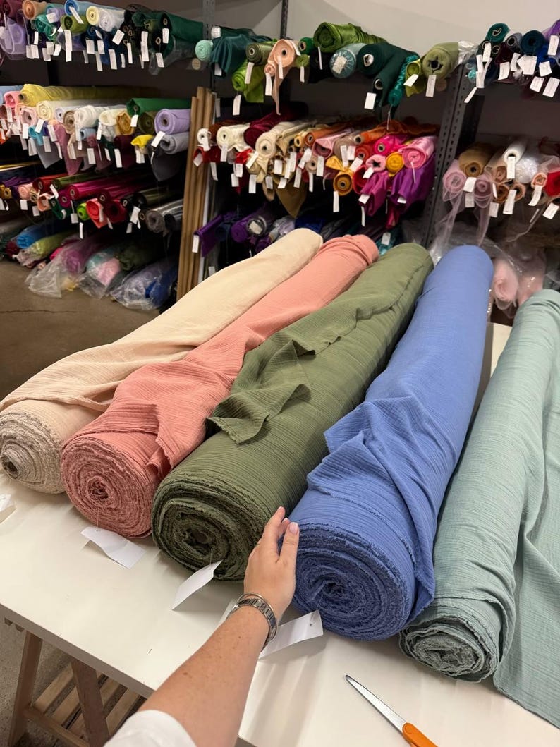 May include: Five large rolls of textured fabric in beige, coral, olive green, blue, and sage green are arranged on a white table. Behind the table, metal shelving units are filled with numerous additional rolls of fabric in various colors.