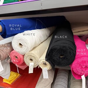 May include: Multiple fabric rolls stored on a shelf, labeled with colors including royal blue, white, black, hot pink, earthy brown, coral, oat, and raw linen. Each roll is cylindrical with a cardboard core, showing various textures and shades.