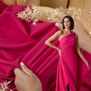 May include: A bright pink fabric with a woman wearing a one-shoulder pink dress with a slit up the leg. The fabric is draped over a hand and the woman is holding the fabric in her other hand. The text "SECRET SPARK fabrics" is visible in the top center of the image.