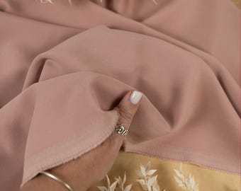 May include: A close-up of a dusty rose colored fabric. The fabric is soft and smooth, and it has a subtle sheen. The fabric is being held by a hand, and the edge of the fabric is visible. The fabric is on a light brown surface.