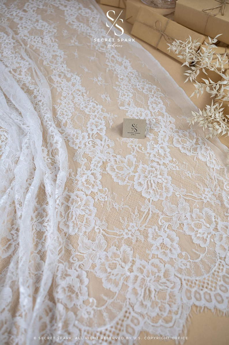 May include: White floral lace fabric with a delicate pattern. The fabric is draped over a neutral surface, showcasing the intricate details of the lace. The image includes the text "SECRET SPARK fabrics".