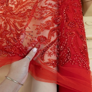 Puede incluir: Primer plano de tela roja con intrincados bordados florales y abalorios. La tela está superpuesta a un material transparente de color nude. El texto "SECRET SPARK" es visible en la parte superior. Información de derechos de autor en la parte inferior.