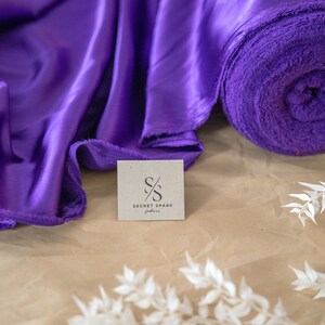 Pu&ograve; includere: Tessuto di raso viola e un rotolo di tessuto viola con un'etichetta di carta marrone che dice "Secret Spark Fabrics" con un logo stilizzato "S/S".