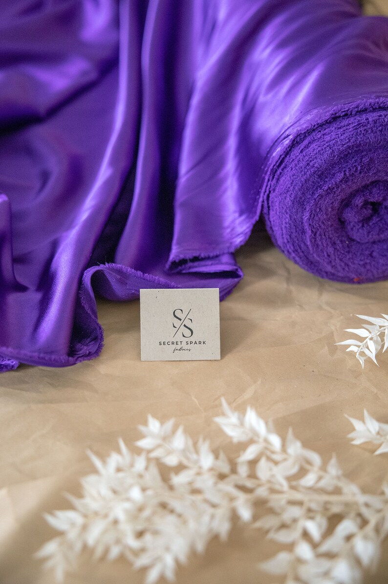 Pu&ograve; includere: Tessuto di raso viola e un rotolo di tessuto viola con un'etichetta di carta marrone che dice "Secret Spark Fabrics" con un logo stilizzato "S/S".