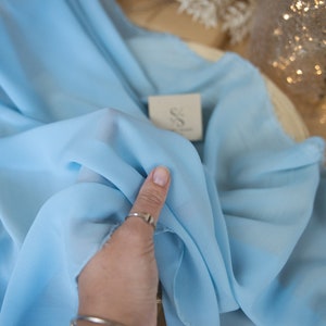 May include: Light blue fabric with a soft, flowing texture. The fabric is held up, revealing its delicate nature. A small tag with the text "SECRET SPARK fabrics" is visible. The image is set against a neutral background.