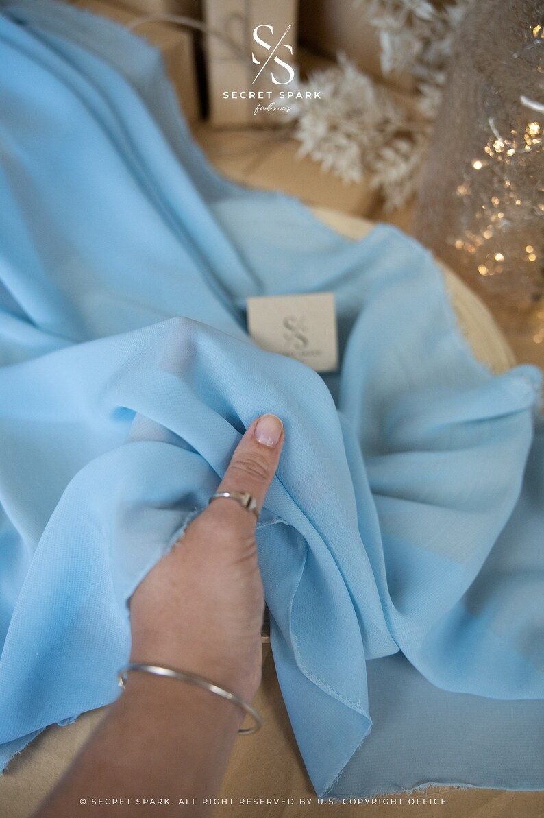 May include: Light blue fabric with a soft, flowing texture. The fabric is held up, revealing its delicate nature. A small tag with the text "SECRET SPARK fabrics" is visible. The image is set against a neutral background.