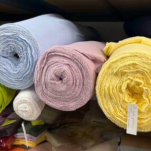 May include: Multiple large rolls of fabric in various colors including light blue, soft pink, bright yellow, lime green, and white. The yellow roll features a white paper tag with handwritten text that reads Soft yellow 100% cotton.