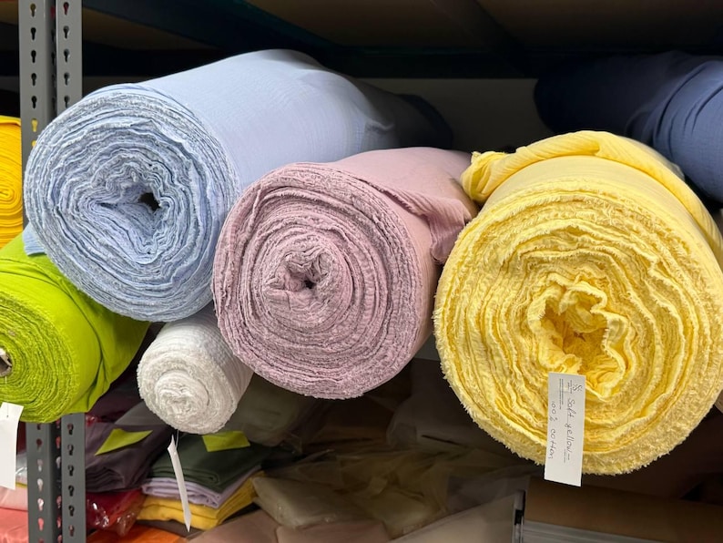 May include: Multiple large rolls of fabric in various colors including light blue, soft pink, bright yellow, lime green, and white. The yellow roll features a white paper tag with handwritten text that reads Soft yellow 100% cotton.