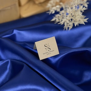 Może przedstawiać: Brązowa karta z tekstem "SECRET SPARK fabrics" i logo "S/S" na tle niebieskiego satynowego materiału.