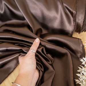 May include: A close-up of a smooth, dark brown satin fabric. The fabric is draped over a surface and a hand is holding a corner of the fabric. The fabric is soft and luxurious.