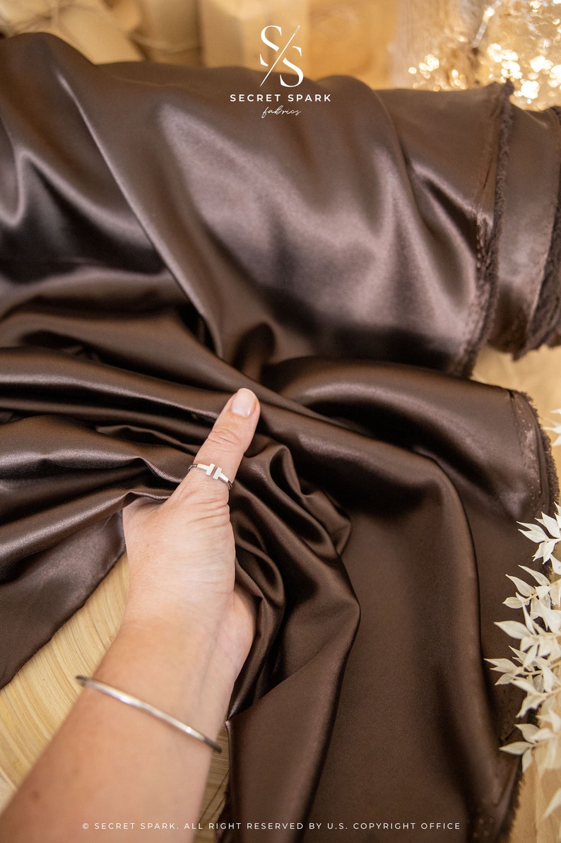 May include: A close-up of a smooth, dark brown satin fabric. The fabric is draped over a surface and a hand is holding a corner of the fabric. The fabric is soft and luxurious.