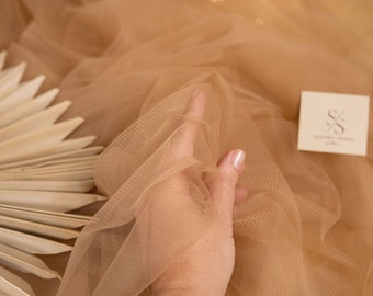 May include: A close-up of a hand holding a piece of sheer, light brown tulle fabric. The fabric is soft and delicate, with a subtle shimmer. The background is blurred and out of focus, with a hint of golden light.