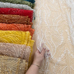 May include: A selection of beaded fabric swatches in shades of blue, green, silver, red, orange, yellow, and gold. A hand holds a piece of white fabric with a leaf design. The text "SECRET SPARK" is visible.