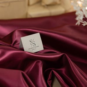 May include: A close-up of a burgundy satin fabric with a small white card on top. The card has a black logo with the text "SECRET SPARK fabrics".