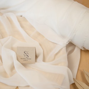 K&ouml;nnte beinhalten: Ein wei&szlig;er, durchsichtiger Stoff mit ausgefransten Kanten. Der Stoff ist auf einer Holzoberfl&auml;che gefaltet und drapiert. Eine kleine braune Karte mit dem Text "SECRET SPARK fabrics" ist in der Mitte des Bildes zu sehen.