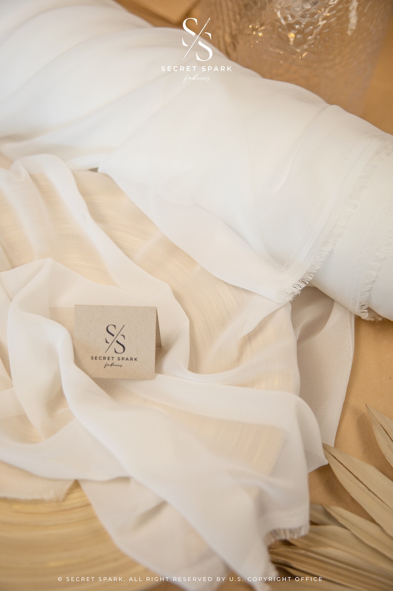 K&ouml;nnte beinhalten: Ein wei&szlig;er, durchsichtiger Stoff mit ausgefransten Kanten. Der Stoff ist auf einer Holzoberfl&auml;che gefaltet und drapiert. Eine kleine braune Karte mit dem Text "SECRET SPARK fabrics" ist in der Mitte des Bildes zu sehen.