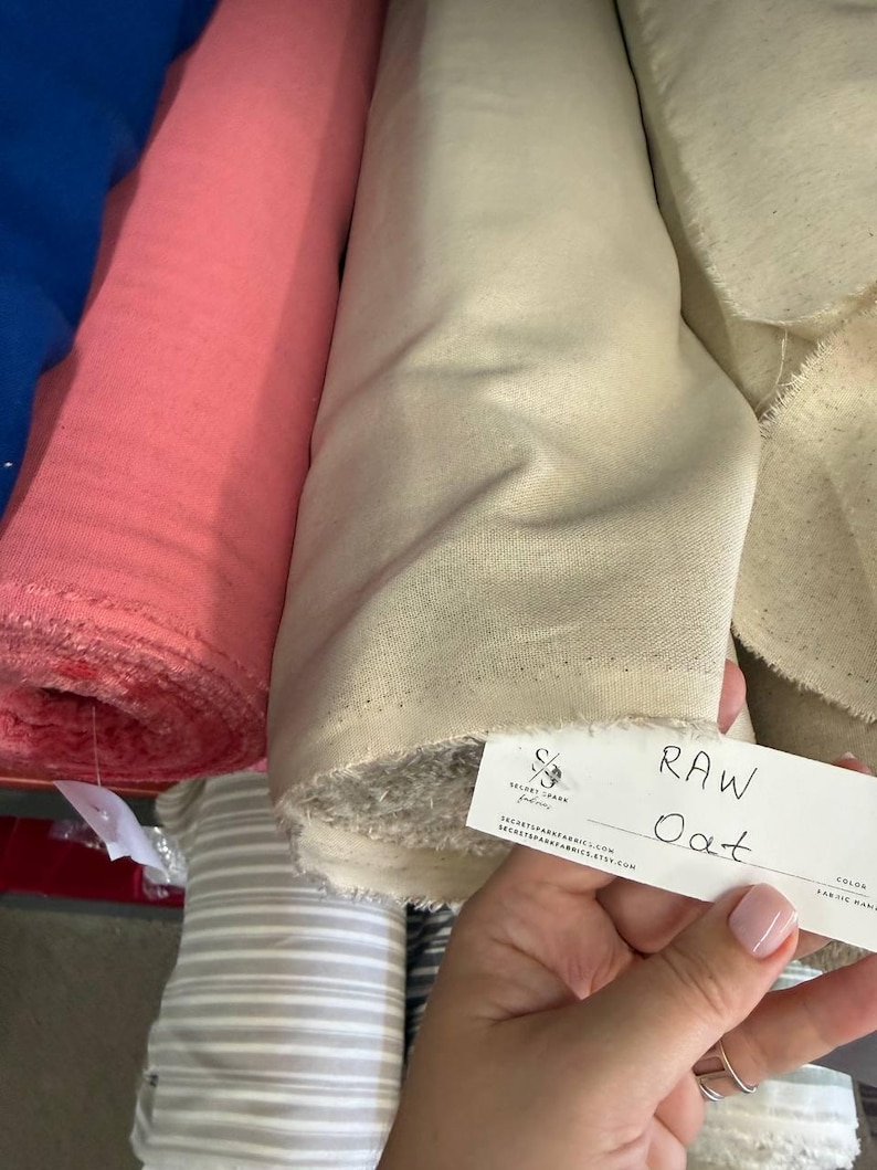 May include: Several rolls of fabric in various colors including blue, pink, and a light beige labeled Raw Oat. The beige fabric roll features a white tag with the text Secret Spark Fabrics, the website URL, and the handwritten color name Raw Oat.