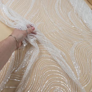 May include: White bridal fabric with a flowing, wave-like pattern. The fabric is embellished with sequins and beads, creating a shimmering effect. The Secret Spark Fabrics logo is visible in the upper left corner.