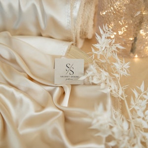 Könnte beinhalten: Nahaufnahme eines beigefarbenen Satin-Stoffes mit einer kleinen braunen Karte mit dem Logo "SECRET SPARK fabrics".