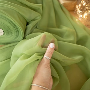 May include: A roll of light green sheer fabric, also known as chiffon, is shown with a hand holding a piece of the fabric. The fabric is soft and flowing. The text "SECRET SPARK fabrics" is visible in the image.