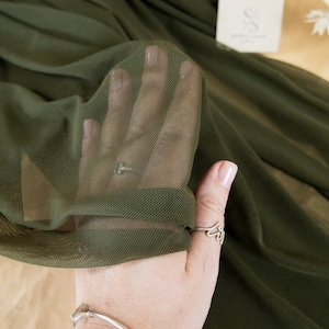 May include: Close-up of a green mesh fabric. The fabric is held in a hand with a silver ring and bracelet. The fabric is soft and sheer, with a fine weave. The text "SECRET SPARK fabrics" is visible in the top left corner of the image.