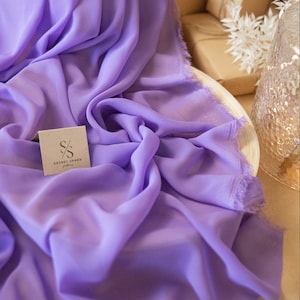 Könnte beinhalten: Ein helllilafarbener, durchsichtiger Stoff mit ausgefransten Kanten. Der Stoff ist gefaltet und auf einer Holzoberfläche drapiert. Eine kleine weiße Karte mit dem Text "SECRET SPARK fabrics" ist in der linken unteren Ecke des Bildes zu sehen.