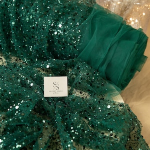 May include: Emerald green sequin fabric with a sheer mesh backing. The fabric is rolled up and has a small white tag with the text "SECRET SPARK fabrics" on it.