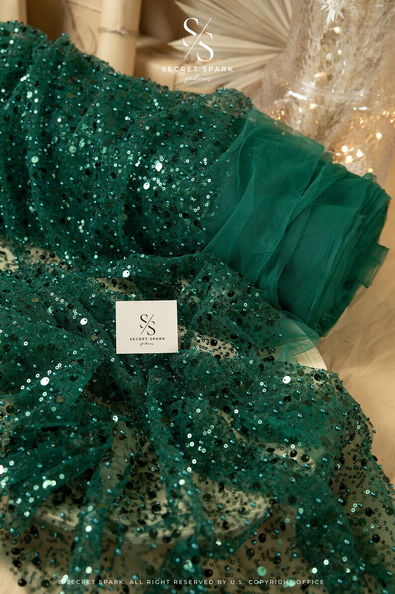 May include: Emerald green sequin fabric with a sheer mesh backing. The fabric is rolled up and has a small white tag with the text "SECRET SPARK fabrics" on it.