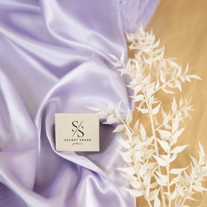 May include: A close-up of a light purple satin fabric with a small white card that says "SECRET SPARK fabrics" on it. A white dried flower branch is on the right side of the image.
