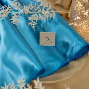 Puede incluir: Un primer plano de una tela de satén turquesa brillante doblada sobre una superficie de madera. La tela es lisa y brillante, con un brillo sutil. La tela está etiquetada con una pequeña etiqueta marrón que dice "SECRET SPARK fabrics".