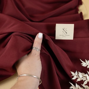 May include: A close-up of a deep burgundy fabric with a soft, smooth texture. The fabric is draped over a surface and held by a hand wearing a silver ring. The fabric is labelled "SECRET SPARK fabrics".