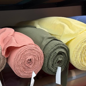 May include: Four rolls of textured fabric in shades of light pink, coral, olive green, and pale yellow are arranged side by side on a shelf. Each roll features a crinkled surface and includes a small white paper tag attached to the end.