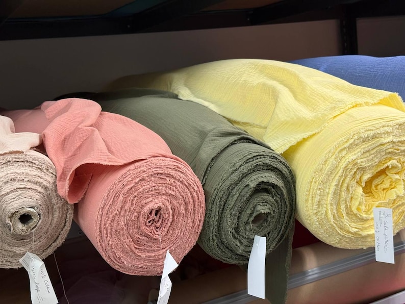 May include: Four rolls of textured fabric in shades of light pink, coral, olive green, and pale yellow are arranged side by side on a shelf. Each roll features a crinkled surface and includes a small white paper tag attached to the end.