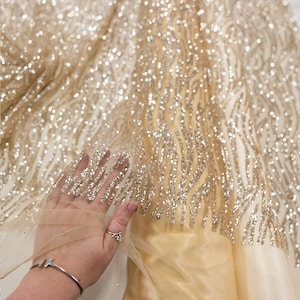 Pode incluir: Close-up de um tecido brilhante com um design de lantejoulas douradas e prateadas. O tecido tem um padrão fluido e ondulado. O texto "SECRET SPARK fabrics" é visível.